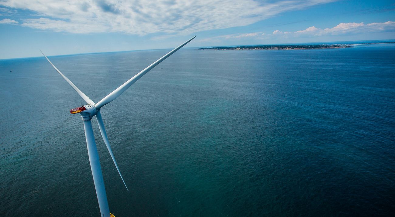 A wind turbine is pictured at left and sits on top of a wide open ocean, with a small patch of land with green grass in view. Clouds are seen on a blue sky.