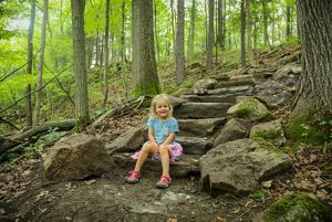 A small child smiles and sits on the bottom step of a rock stairway in the woods.
