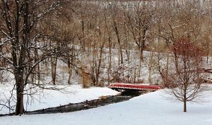 A narrow creek bends between snow covered banks. A red foot bridge spans the water. Tall leafless trees grow along the sides of the creek.