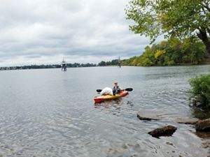 A woman wearing a PA DCNR uniform and cap floats in a kayak in a wide marsh. There is a large white bag of trash balanced on the bow of the orange kayak, collected during a volunteer event.