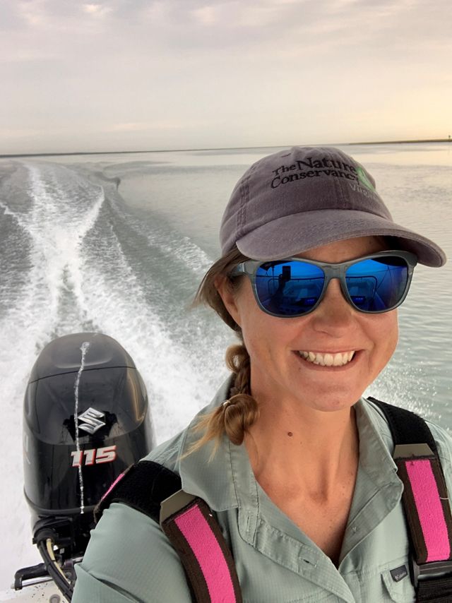 Candid snapshot of Marine Restoration Specialist Brittany Gonzalez. A smiling woman wearing sunglasses and a ball cap drives a small boat in open water.