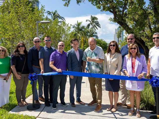 A group of people stand behind a ribbon as one man with a large scissors cuts the blue ribbon.