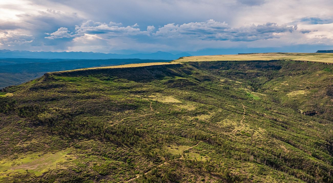 Aerial view of a vast forested mountain landscape and a flat mesa with a dirt trail running through it.