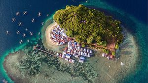 Vista aérea de pequena ilha tropical com vegetação densa, vila costeira, barcos no mar azul-turquesa e recifes visíveis ao redor.