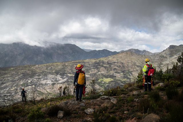 People with gear standing atop a mount overlooking rolling mountain peaks with their backs to the camera.