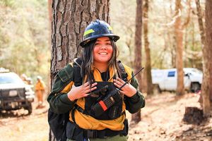 A woman in firefighting gear stands in front of a brown tree trunk.