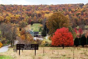 A large wooden sign welcomes visitors to Cherry Valley National Wildlife Refuge. A tree with bright red leaves stands in an open field to the right. A forest of fall colors dominates the background.