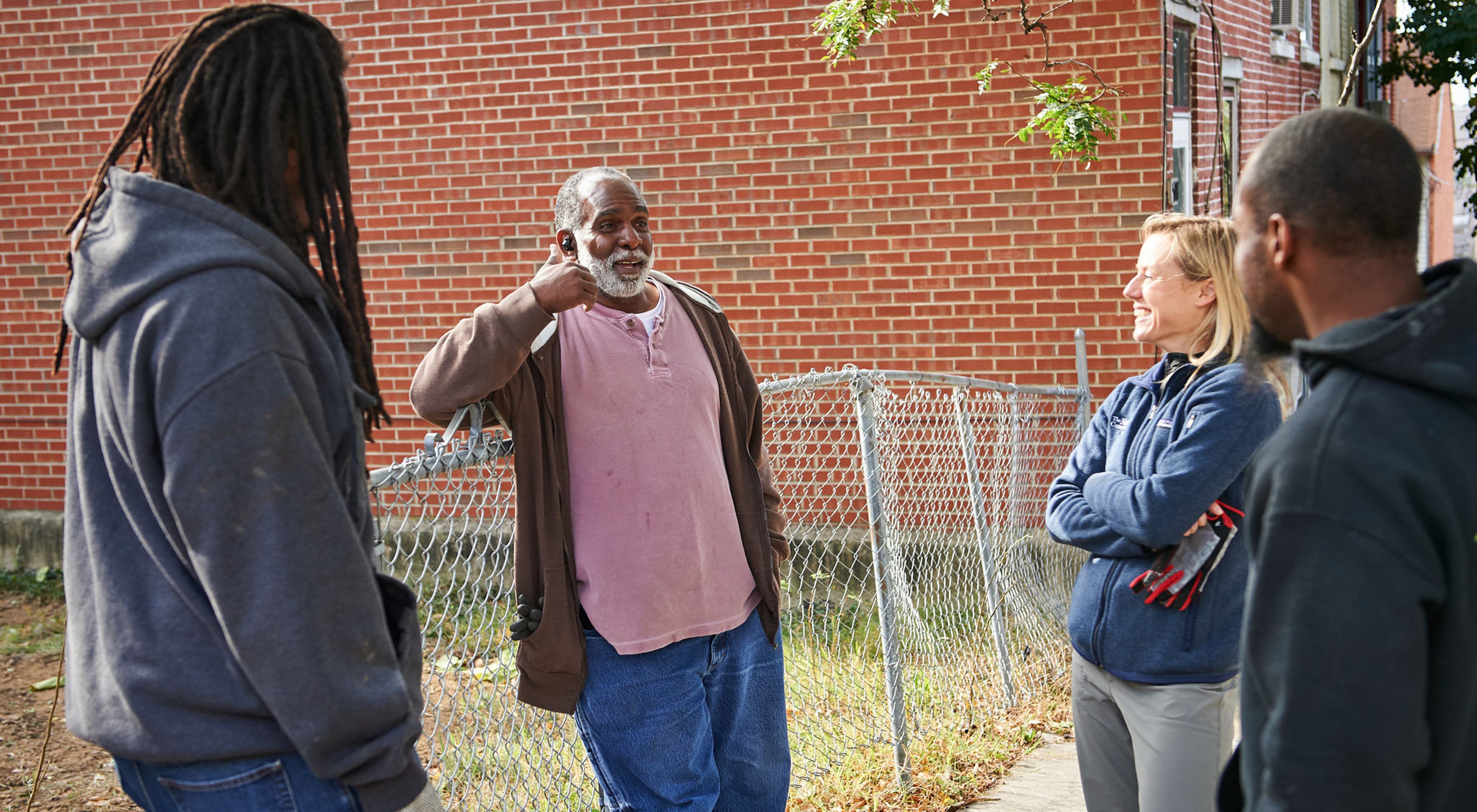 Four people stand together talking next to an open lot. The man in the center is the focus of attention. He is smiling and gesturing as he talks.