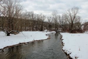 A wide creek flows between snow covered banks. A mountain ridge is visible in the background behind a row of tall leafless trees. Heavy clouds hang low in the sky.