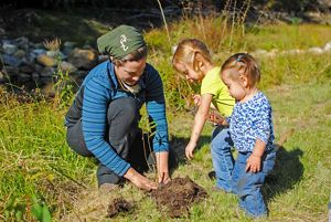 2 niños pequeños observan cómo un voluntario siembra una planta en una zona de pasto cerca de un río.