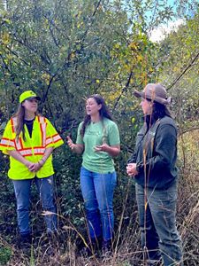 Three people stand in a natural setting with tall grass and bushes. One is wearing a bright yellow safety vest and cap. Another is gesturing while speaking, clad in a green T-shirt and jeans. The third person is dressed in a dark jacket and hat. They seem to be engaged in conversation outdoors.