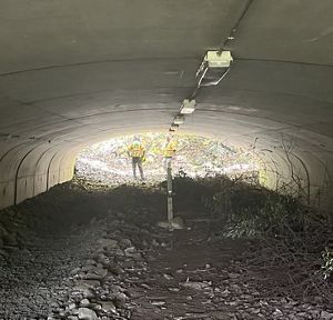 A person wearing a safety vest is standing in a large, concrete tunnel, holding surveying equipment. The tunnel is lined with cables and opens up to daylight, with foliage visible at the far end.