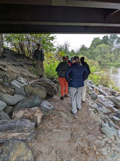 People walk on a path made of dirt and stone that runs alongside a stream, all of which is within a culvert under a roadway.