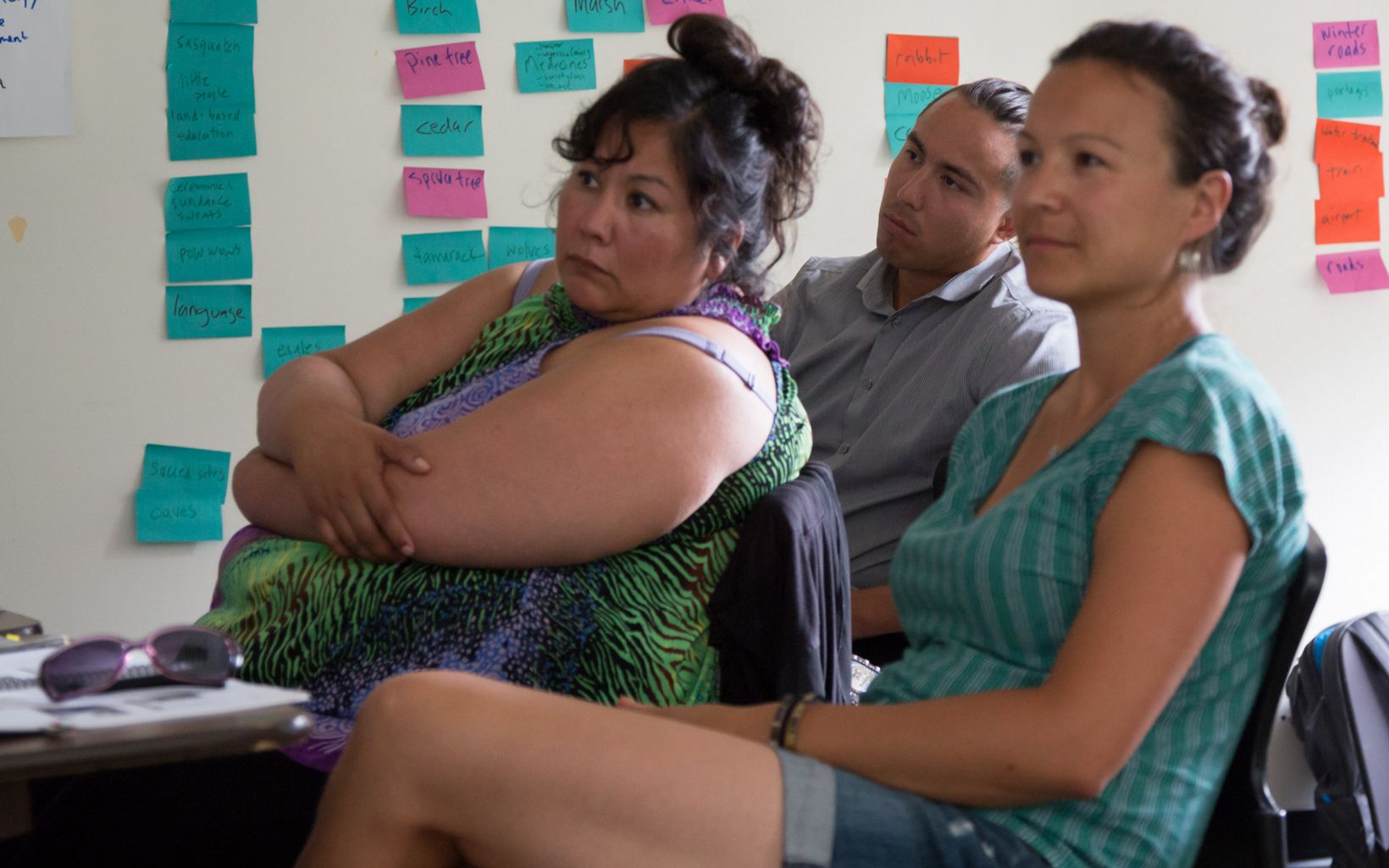 Three people sit in a classroom; a white board with colorful sticky notes is in the background.