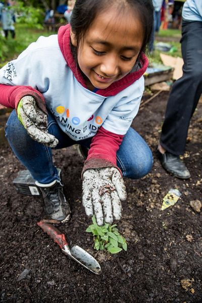 A child crouches in the dirt at a school garden. She holds her gloved hand out to show a small earthworm curled up in her palm.