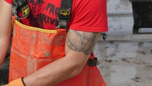 Cropped view of a man shucking an oyster. He is wearing orange overalls. A large oyster tattoo is visible on his bicep.