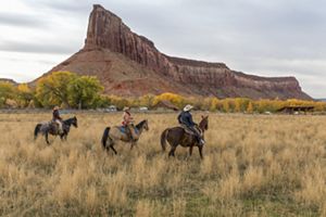Three people in jeans and western hats ride horses in high grass. A red rock formation juts sharply upward in the distance.