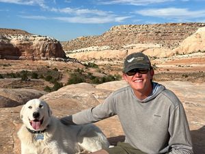A man and his dog sitting down facing camera with mountain landscape in background.