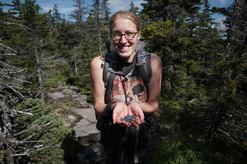 A smiling woman holds freshly picked wild blueberries in her cupped hands. She is standing on a trail surrounded by tall trees.