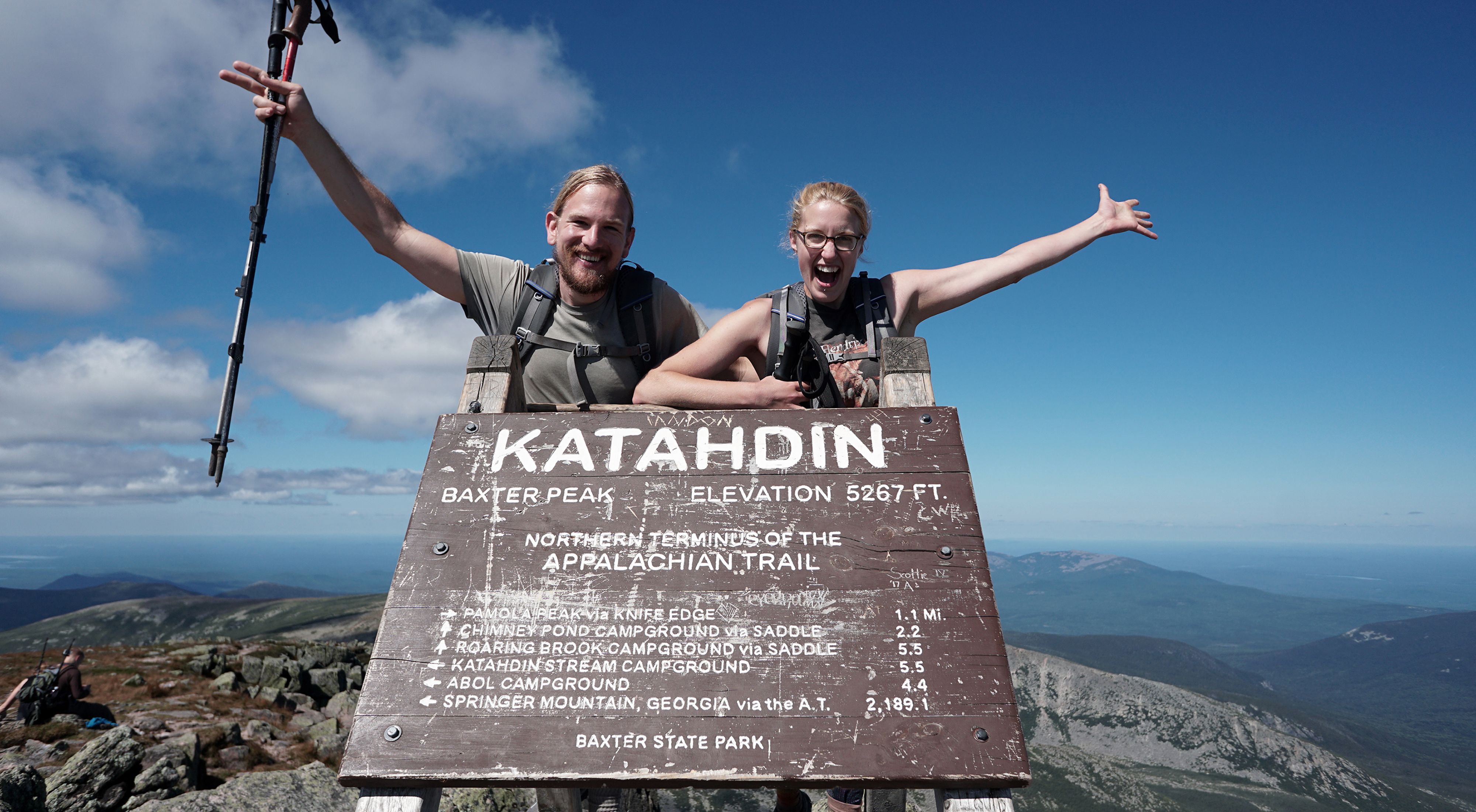 A man and woman stretch out their arms in celebration. They're standing behind a large sign marked KATAHDIN that marks the northern terminus of the Appalachian Trail.
