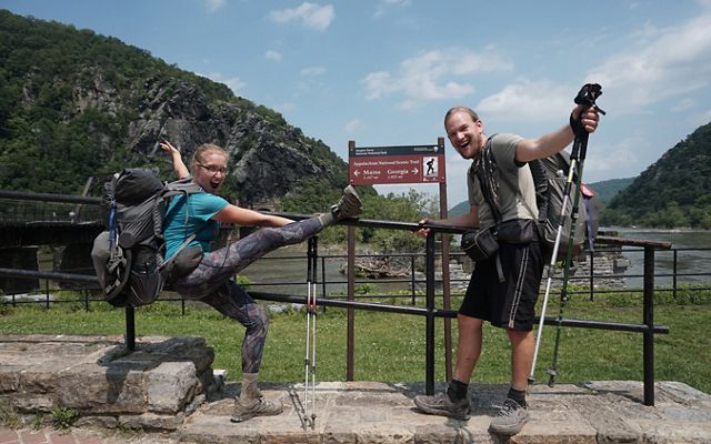 A woman and man celebrate and pose next to a sign marking the midpoint of the Appalachian Trail in Harpers Ferry, WV.
