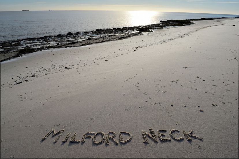 A wide open stretch of beach ends at a shoreline edged with seaweed. The sun reflects on the calm surface of the Delaware Bay. In the foreground the words, Milford Neck, have been written in the sand.
