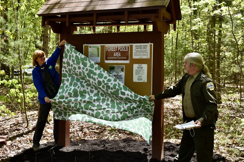 Two people remove a green and white sheet from a new preserve kiosk, part of a land transfer ceremony with the state of Pennsylvania. The kiosk contains signs and visitor information.