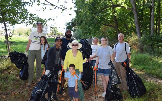 A group of nine people pose together for a photo during a clean up event. The diverse group is holding large black trash bags and bright orange grabbers.