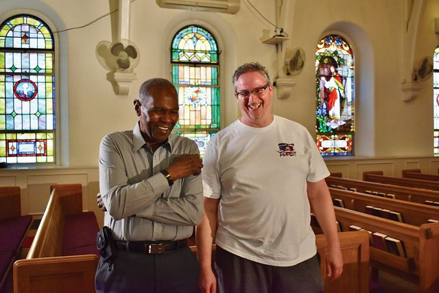 Two men stand together in a church sanctuary. Rows of wooden pews and colorful stained glass windows are behind them.