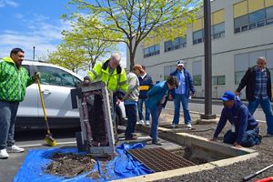 One person holds a drainage cover in a parking lot while a group of people bend over to see inside.