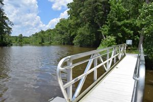 View from the end of a narrow floating dock with high metal sides. The dock leads to a path that disappears into a thick forest. Trees line the banks of the wide river that stretches to the horizon.