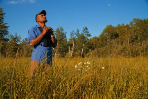 A man stands in a grassy field while holding binoculars and looking toward the sky.