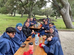 Students wearing rain ponchos sit at picnic tables while having a snack.