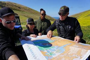 A group of scientists look at a map, standing outside in a rolling, grassy field with ocean in the distance.