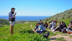 A man stands and speaks in front of a group of sitting students in a grassy meadow with ocean in the background.