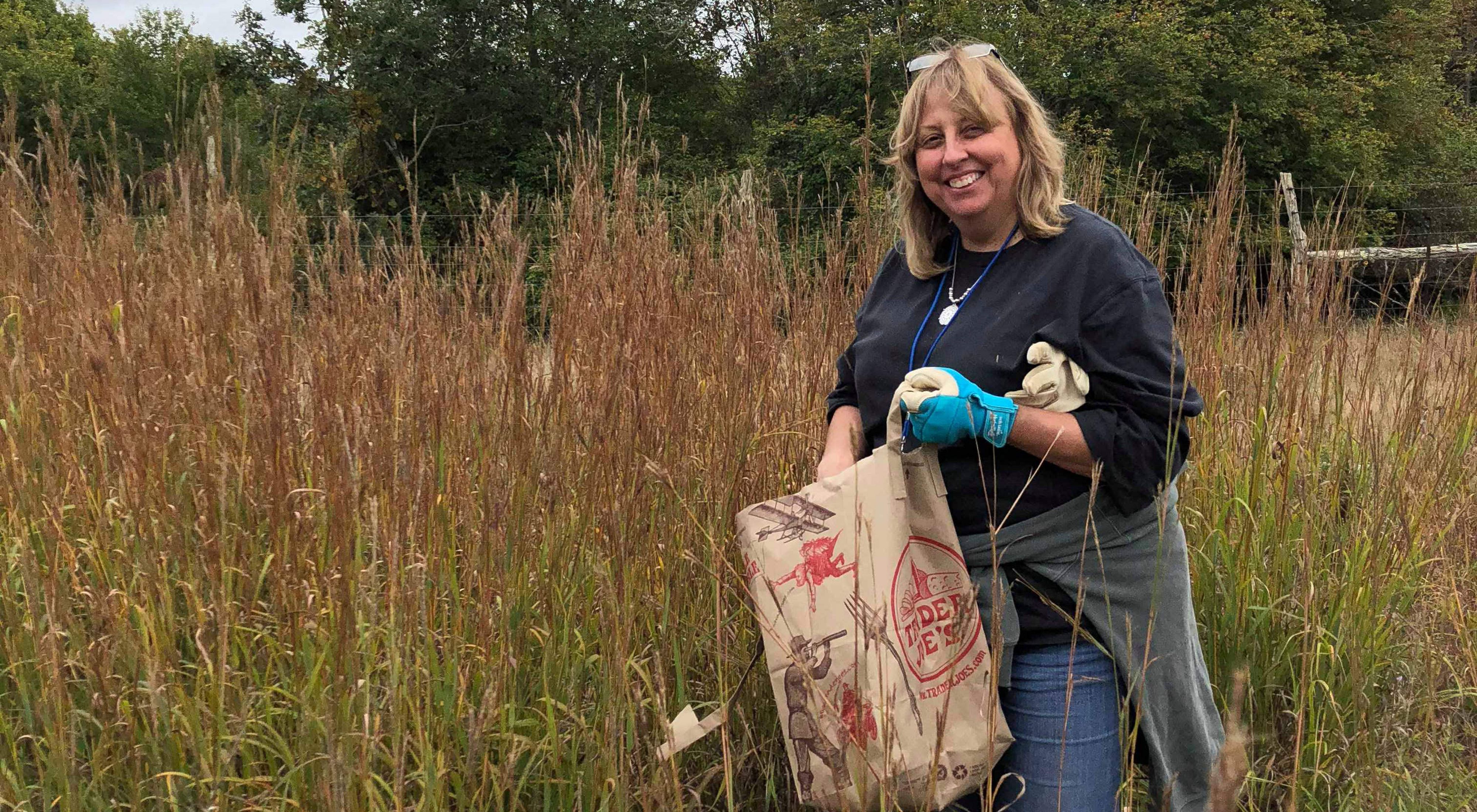 A smiling volunteer wearing gardening gloves puts trash in a paper bag.