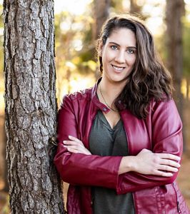 Elizabeth Carter headshot. A smiling, dark haired woman wearing a gray top and red leather jacket leans again a tree.