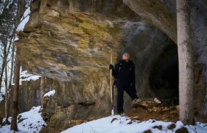 A person dressed in winter clothing, holding a hiking stick, stands next to a large rock outcropping in the snow.