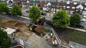 A drone photo looking down onto a parking lot with large yellow construction vehicles and workers digging a hole into brown dirt.