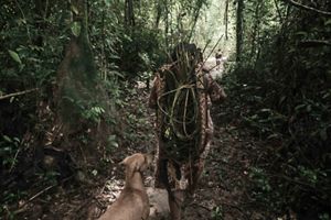 Indígena caminhando na floresta com ferramentas tradicionais.