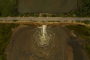 a cracked road stretching over a creek is seen from above with water spilling out of a narrow hole on one side