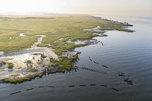a coastline seen from above: homes in the distance, green plants stretching to the horizon, sand and dunes along the water and oyster blocks peeking out of the water offshore