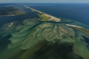 seen from above: a narrow stretch of land surrounded by blue ocean water on both sides is shown with a cut in the middle of the land where the water has pushed through, mud/sand visible on the water