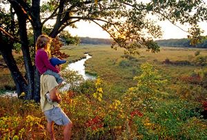 Father with daughter on shoulders looks across wetlands and winding river to forests beyond as setting sun makes fall colors of vegetation glow.