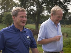 Two men walk down an unpaved farm road. They are both smiling. A line of leafy green trees frame an open field behind them.