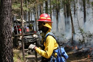 Profile view of a man resting during a controlled burn. He is wearing yellow fire gear and a red hard hat. His hands rest on the handle of a shovel. A low fire burns behind him along a fire line.