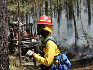 Profile view of a man resting during a controlled burn. He is wearing yellow fire gear and a red hard hat. His hands rest on the handle of a shovel. A low fire burns behind him along a fire line.