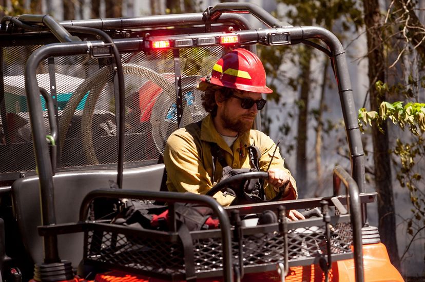 A man wearing a yellow shirt and red hard hat drives a small fire truck. The open sided all terrain vehicle contains hose and water tanks.