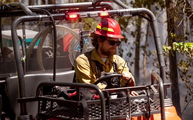 A man wearing a yellow shirt and red hard hat drives a small fire truck. The open sided all terrain vehicle contains hose and water tanks.