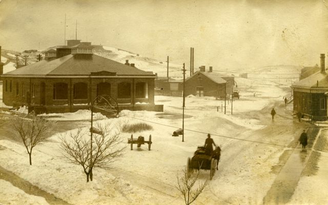 Historical photo of a town square like setting at Army base Fort Terry, with snow on the ground and a horse drawn carriage in foreground. 